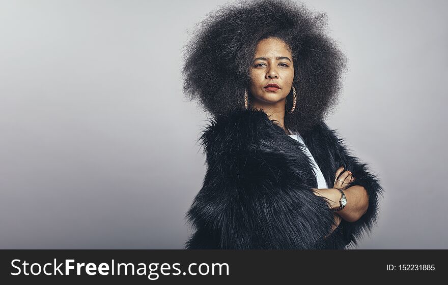 Portrait of woman in fur coat standing with her arms crossed looking at camera. Woman in afro hairstyle standing against grey background