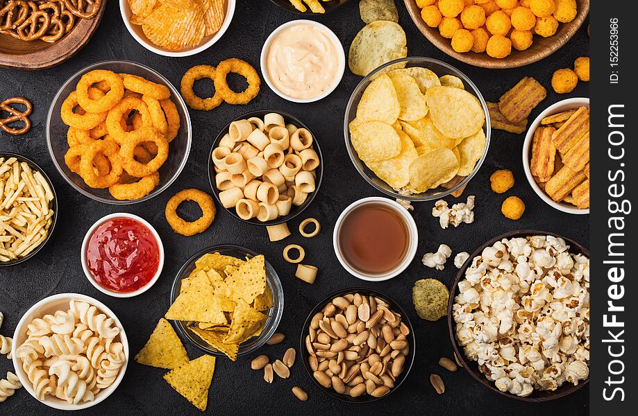 All classic potato snacks with peanuts, popcorn and onion rings and salted pretzels in bowl plates on black background. Twirls