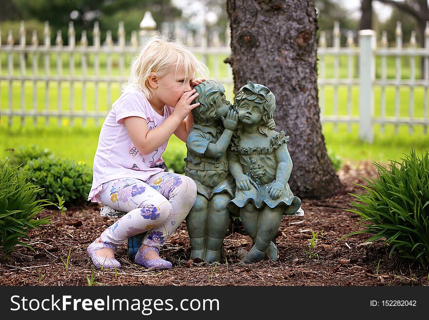 Cute Little Girl Whispering Secret to Little Garden Statue Friends