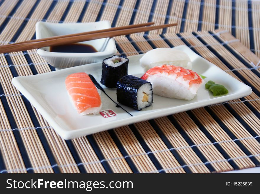 Assortment of sushi on a plate with wood chopsticks and wasabi in a rectangular decorated plate, on a wood table-cloth with soy sauce. Assortment of sushi on a plate with wood chopsticks and wasabi in a rectangular decorated plate, on a wood table-cloth with soy sauce