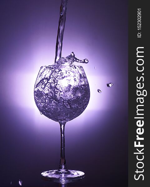 Motion Of Pouring Splash Water In Transparent Wine Glass With Reflect Of Violet Light Backdrop.