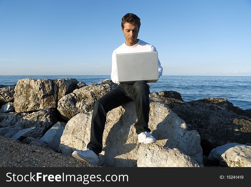 Portrait of young man working with laptop computer sitting outdoor near the shore, blue sky and ocean in background. Portrait of young man working with laptop computer sitting outdoor near the shore, blue sky and ocean in background