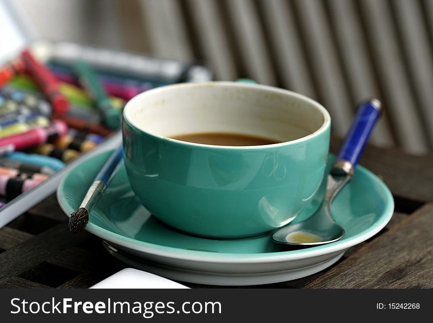 Green coffee cup with a spoon and an artis's painters brush and colorful pens in the background. Green coffee cup with a spoon and an artis's painters brush and colorful pens in the background.