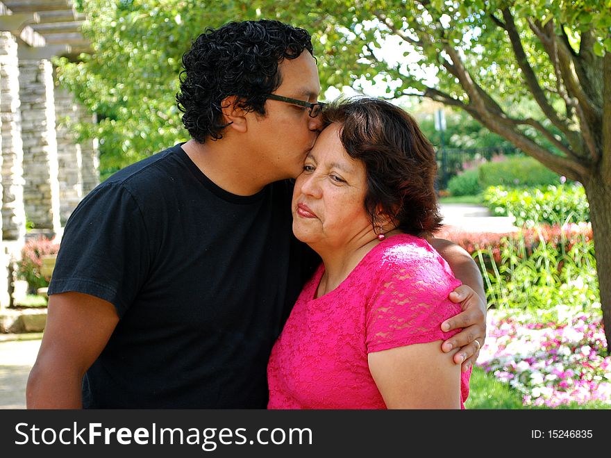 A Hispanic man kisses his mother  at a beautiful park. A Hispanic man kisses his mother  at a beautiful park.