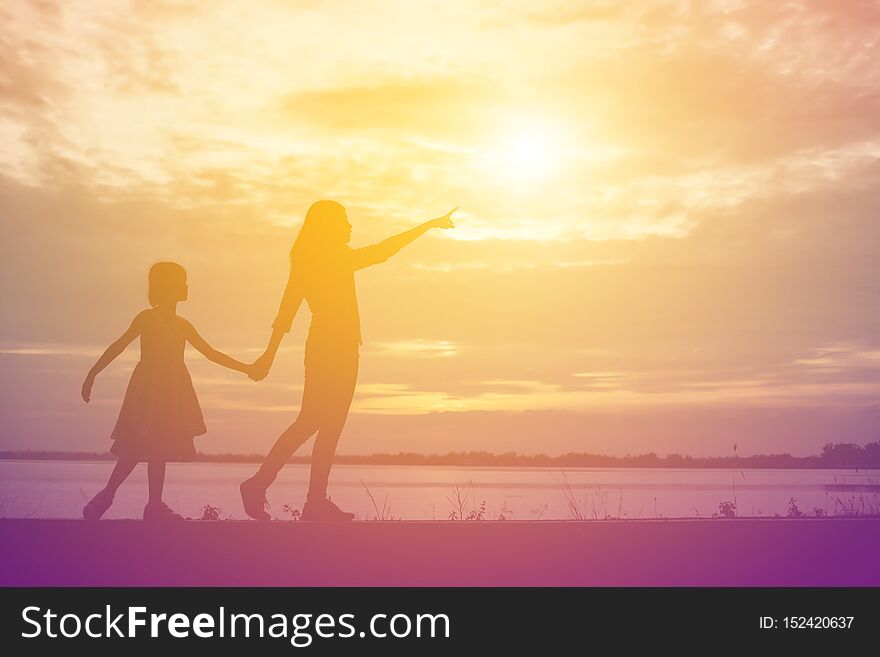 A silhouette of a happy young girl child the arms of his loving mother for a hug, in front of the sunset in the sky on a summer
