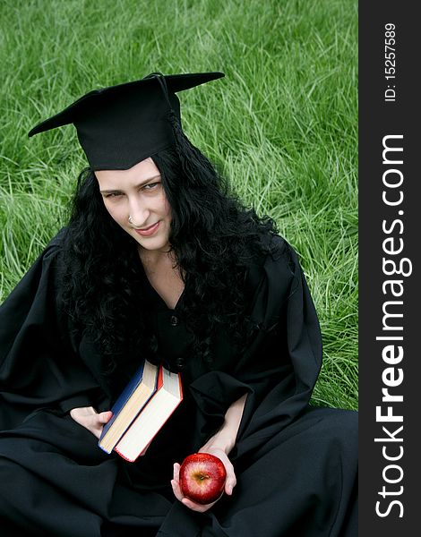 Caucasian student in gown with books sitting on the grass