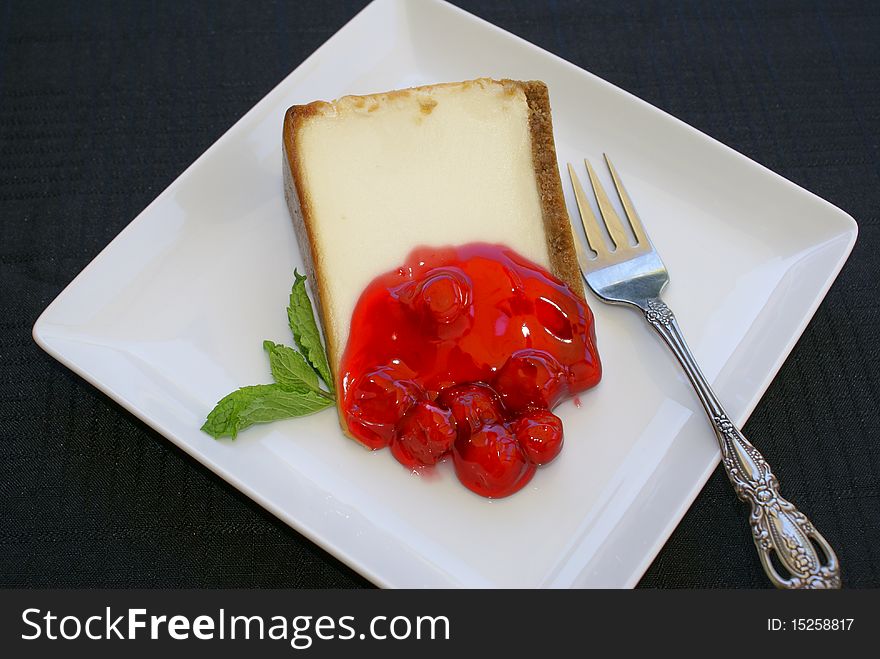 Cheesecake with cherry compote and a sprig of mint on a white plate which is on a black tablecloth with a fork. Cheesecake with cherry compote and a sprig of mint on a white plate which is on a black tablecloth with a fork