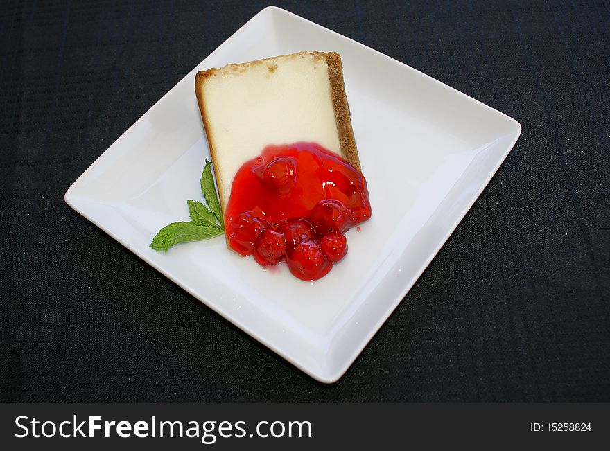 Cheesecake with cherry compote and a sprig of mint on a white plate which is on a black tablecloth. Cheesecake with cherry compote and a sprig of mint on a white plate which is on a black tablecloth