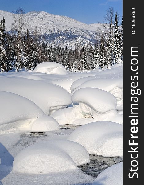 Thick fluffy snowdrifts on the banks and bed of mountain river. Pine forest and further mountain slope on background. Thick fluffy snowdrifts on the banks and bed of mountain river. Pine forest and further mountain slope on background.