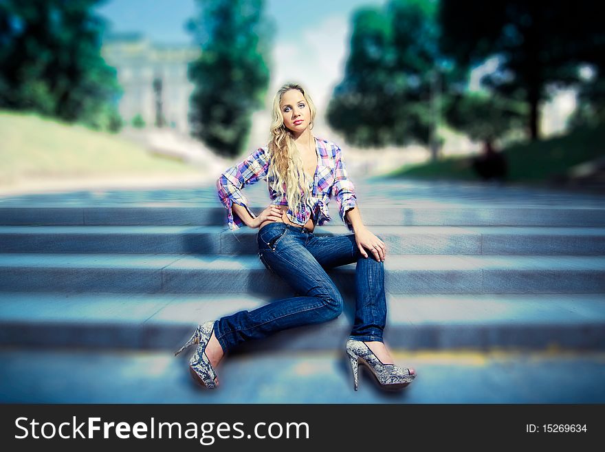 Blonde young woman sitting on stairs, dressed in jeans and a shirt. In the park. Blurred background. Blonde young woman sitting on stairs, dressed in jeans and a shirt. In the park. Blurred background.