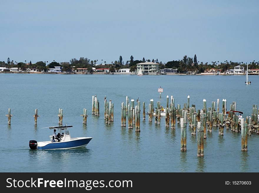 A small fishing boat with two people driving through the bay and pier ruins with pelicans sitting on top. Large houses in the distance on the horizon line, with pale blue sky and water. A small fishing boat with two people driving through the bay and pier ruins with pelicans sitting on top. Large houses in the distance on the horizon line, with pale blue sky and water.