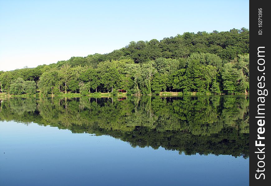 Green trees and a little red structure in the woods reflect vibrantly on a calm river. Green trees and a little red structure in the woods reflect vibrantly on a calm river.
