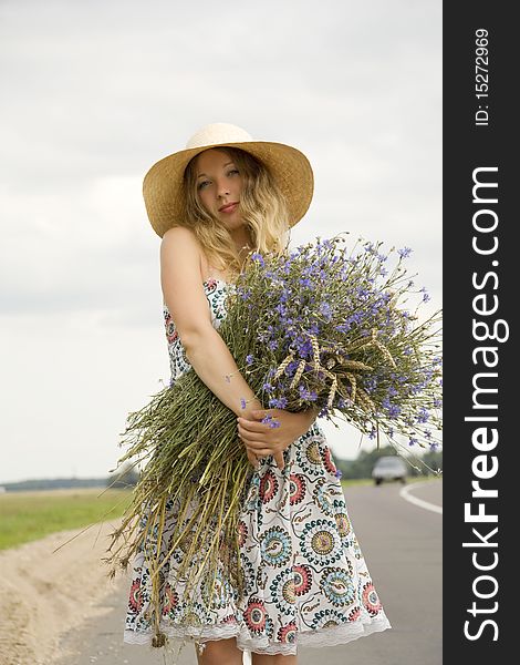 The woman in a hat with a bouquet of cornflowers costs on the brink of road near passing by the car. The woman in a hat with a bouquet of cornflowers costs on the brink of road near passing by the car