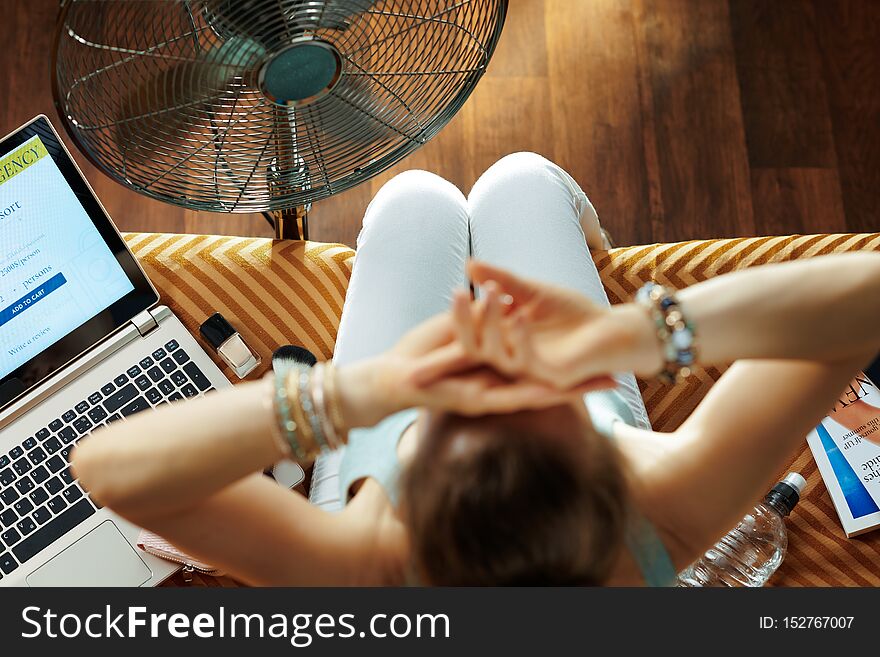 Upper view of relaxed young woman sitting on divan in the modern house in sunny hot summer day using electric floor standing fan. Upper view of relaxed young woman sitting on divan in the modern house in sunny hot summer day using electric floor standing fan