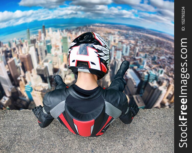 Motorcyclist in full gear and helmet sitting on a tall building. Man looks down on the city