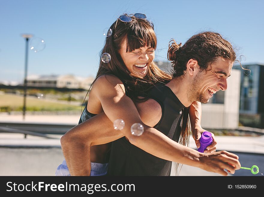 Couple having fun outdoors. Man piggybacking women playing with soap bubble outdoors