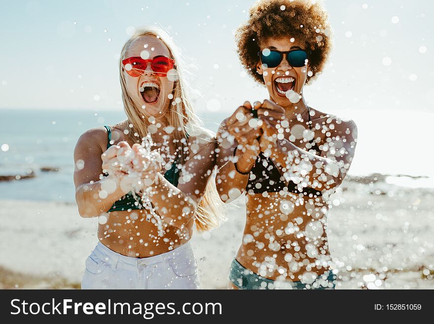 Excited young women friends bursting a water balloon and smiling by the sea. Best friends having fun on beach holiday