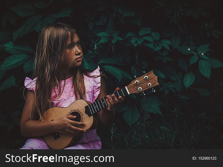 Beautiful young girl with blond hair wearing pink dress playing ukulele and dreaming on dark nature background. Grape leaves. Cutie lady learns play guitar concept. Beautiful young girl with blond hair wearing pink dress playing ukulele and dreaming on dark nature background. Grape leaves. Cutie lady learns play guitar concept