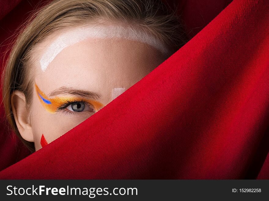 Girl with color make-up on a red background. A blonde with colored make-up peeks out because of the red cloth.