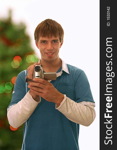 Happy guy holding a camera; blurred christmas tree background. Happy guy holding a camera; blurred christmas tree background