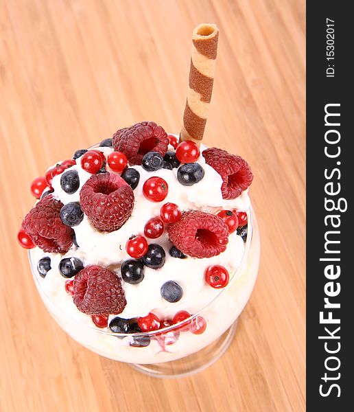 Whipped cream with raspberries, red currants and blue berries in a glass cup, decorated with a wafer tube on wooden background. Whipped cream with raspberries, red currants and blue berries in a glass cup, decorated with a wafer tube on wooden background