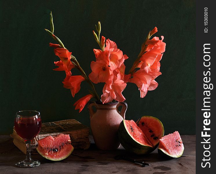 Scarlet gladioluses stand in a vase, the cut water-melon, old books and glass of wine, lies alongside. Scarlet gladioluses stand in a vase, the cut water-melon, old books and glass of wine, lies alongside