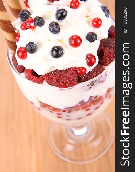 Whipped cream with raspberries, red currants and blue berries in a glass cup, decorated with a wafer tube on wooden background. Whipped cream with raspberries, red currants and blue berries in a glass cup, decorated with a wafer tube on wooden background