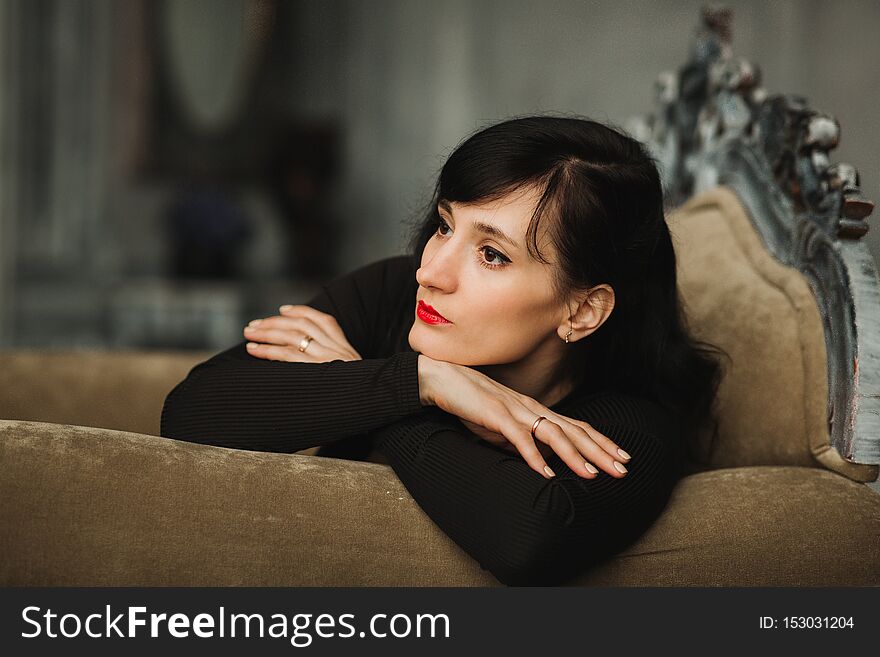 Fashion Portrait Of Young Beautiful Female Model In Black Sexy Dress Sitting On Arm Chair In Interior Studio