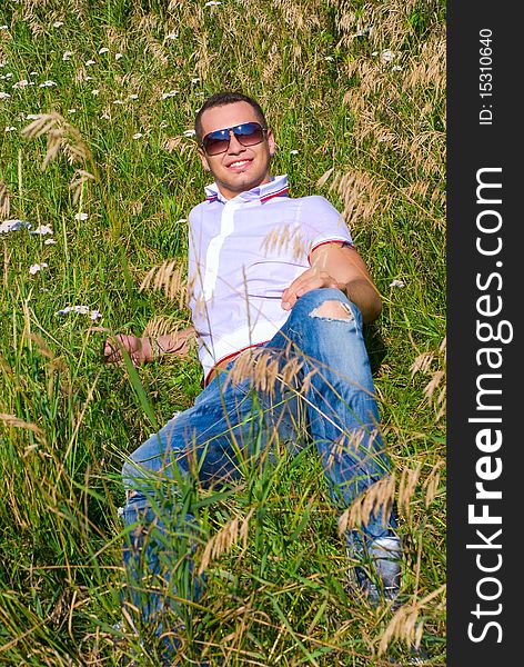 Young man in jeans with teeth smile is laying on summer field. Green grass surrounds him. Young man in jeans with teeth smile is laying on summer field. Green grass surrounds him.