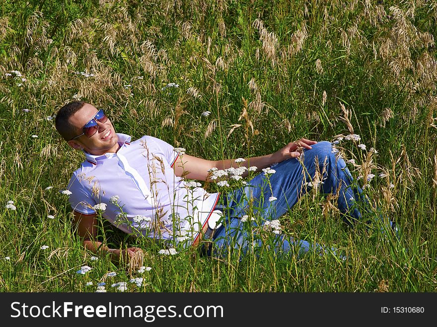 Young man in jeans with teeth smile is laying on summer field. Green grass surrounds him. Young man in jeans with teeth smile is laying on summer field. Green grass surrounds him.