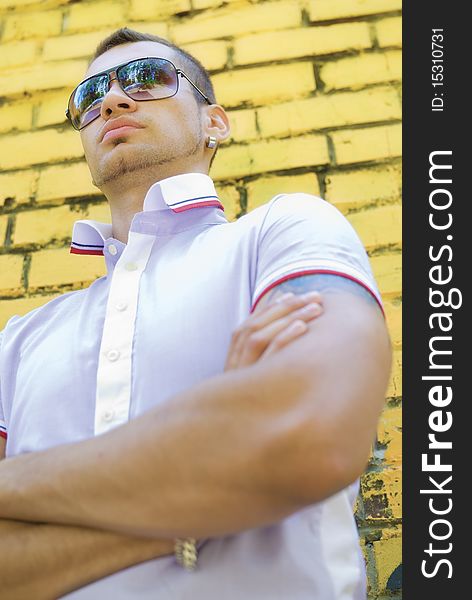 Man with big and muscled arms stands near the yellow brick wall. He wears sun glasses and earrings. Under the shirt you can see part of his tattoo. Man with big and muscled arms stands near the yellow brick wall. He wears sun glasses and earrings. Under the shirt you can see part of his tattoo.