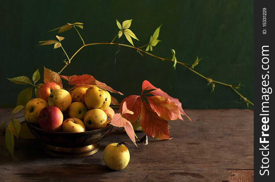 Yellow apples are in a dish, alongside yellow-red leaves. Yellow apples are in a dish, alongside yellow-red leaves