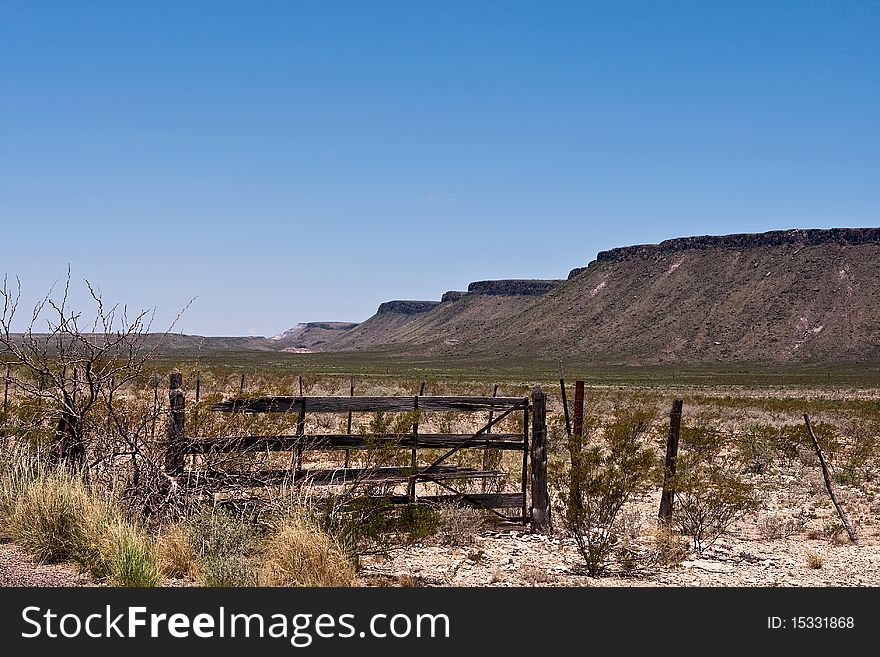 Old wooden gate in desert with mountains in background. Clear blue sky.