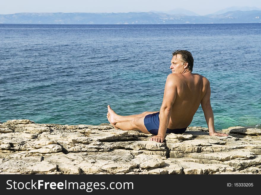 A color photo of a caucasian man sitting on rocks and relaxing next to the stunning blue turquoise sea of Greece. A color photo of a caucasian man sitting on rocks and relaxing next to the stunning blue turquoise sea of Greece.