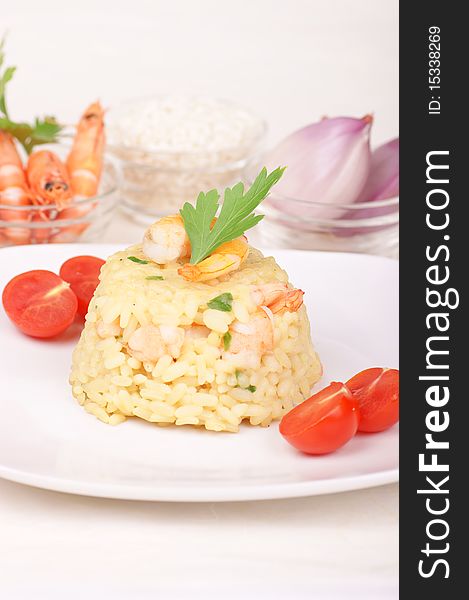 Risotto with shrimps served on a white plate and decorated with cherry tomatoes. Rice, shrimps and shallots in small glass bowls are out of focus in the background. Studio shot. Shallow DOF