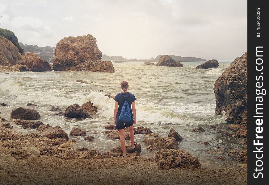 Adventurous girl in shorts with blue backpack looking at stormy sea. Calm view of back photo. Travel the world and explore, be free. Adventurous girl in shorts with blue backpack looking at stormy sea. Calm view of back photo. Travel the world and explore, be free.