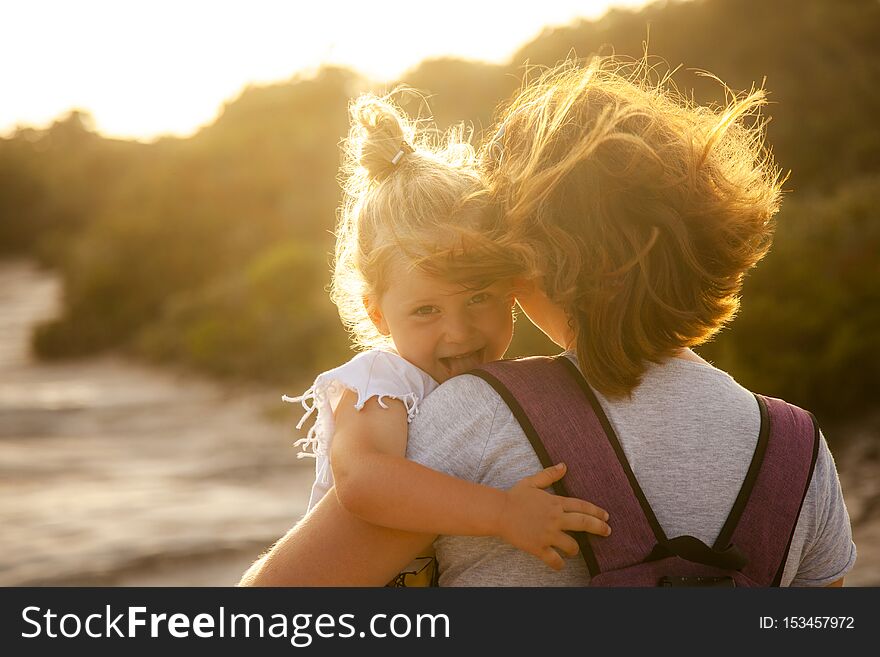 Portrait of a 3 year old Caucasian girl with blond hair who shows her tongue playfully. In the arms of the mother, backlit image at sunset