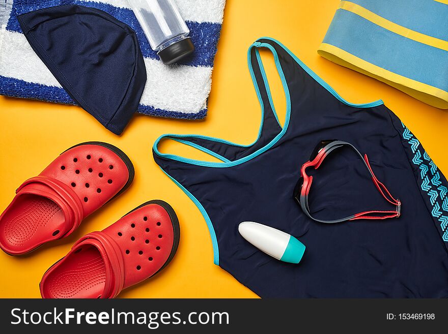 Flat lay summer pool accessories such such as blue swimwear, swimming cap and goggles, striped towel, deodorant, red slippers and transparent bottle on a yellow background. Colorful beach wear. Copy space, top view, close-up. Flat lay summer pool accessories such such as blue swimwear, swimming cap and goggles, striped towel, deodorant, red slippers and transparent bottle on a yellow background. Colorful beach wear. Copy space, top view, close-up.