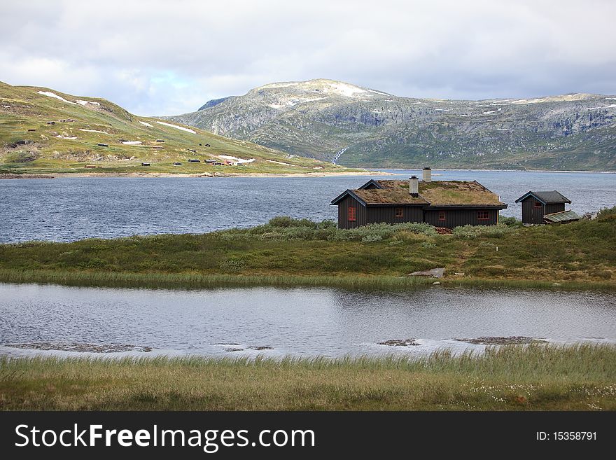 Traditional norwegian house (hytta) placed in the middle of the lake, surrounded by the mountains and the rugged landscape. Grass growing on the roof of the house. Traditional norwegian house (hytta) placed in the middle of the lake, surrounded by the mountains and the rugged landscape. Grass growing on the roof of the house.