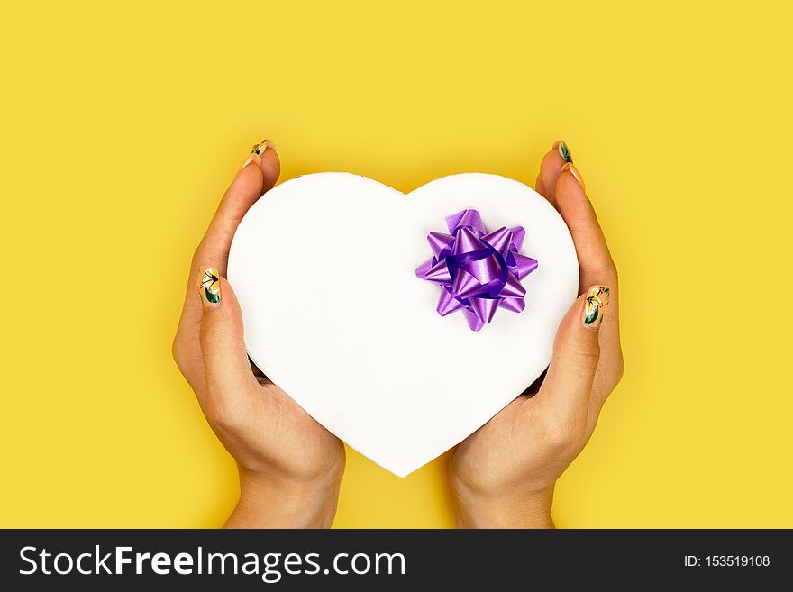 Heart shaped Valentines Day gift box in the hands of the girl on yellow paper background.