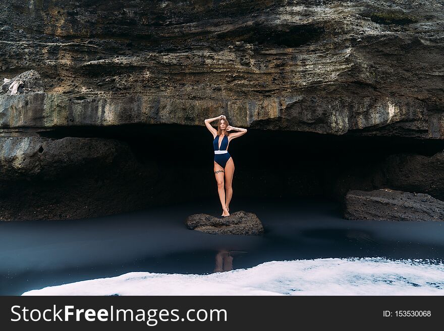 Sexy girl poses in blue swimsuit near cave on black sand beach, concept travel