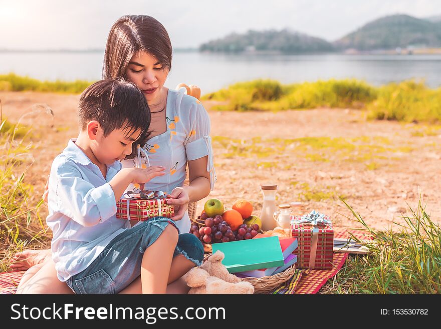 Beautiful Asian mother and son doing picnic and opening gift box from surprise in Birthday party on meadow near lake and mountain