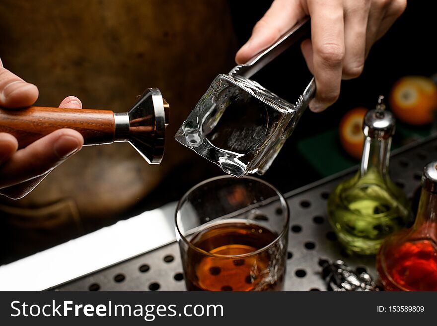 Professional bartender making a stamp imprint on the big ice cube in the glass on the bar counter in the dark blurred background in the dark