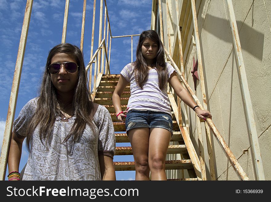 A couple of teenagers, one in sunglasses standing on a metal staircase with a bright blue sky overheard. A couple of teenagers, one in sunglasses standing on a metal staircase with a bright blue sky overheard.