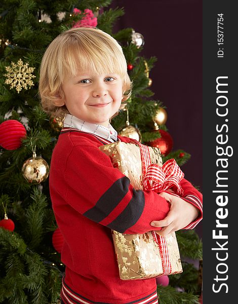 Young Boy Holding Gift In Front Of Christmas Tree Smiling