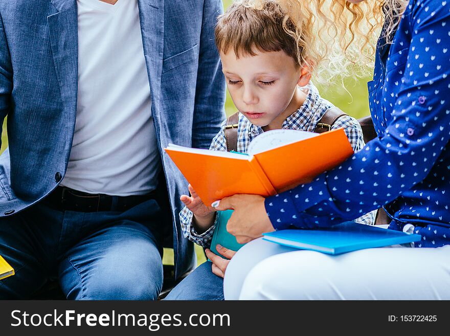 Engaged child boy sitting between mother and father and reading interesting book outdoor.
