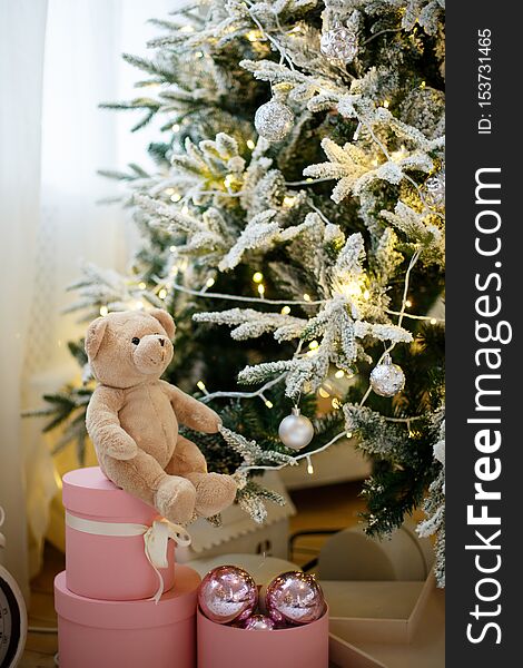 A teddy bear toy is sitting on a pink gift box, at the Christmas tree decorated with yellow garlands and balls. A teddy bear toy is sitting on a pink gift box, at the Christmas tree decorated with yellow garlands and balls