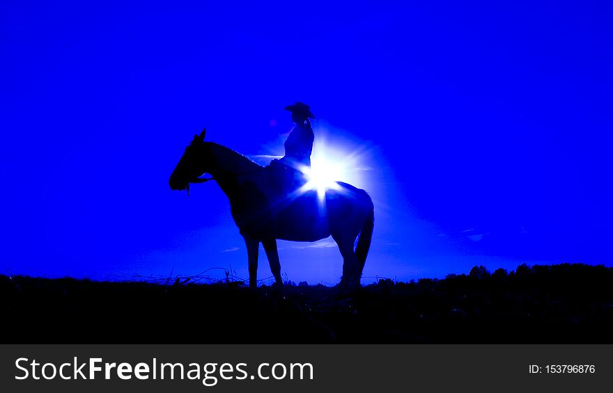 Silhouette of a cowgirl on open plain in western  riding a horse lit by the setting sun in blue. Silhouette of a cowgirl on open plain in western  riding a horse lit by the setting sun in blue