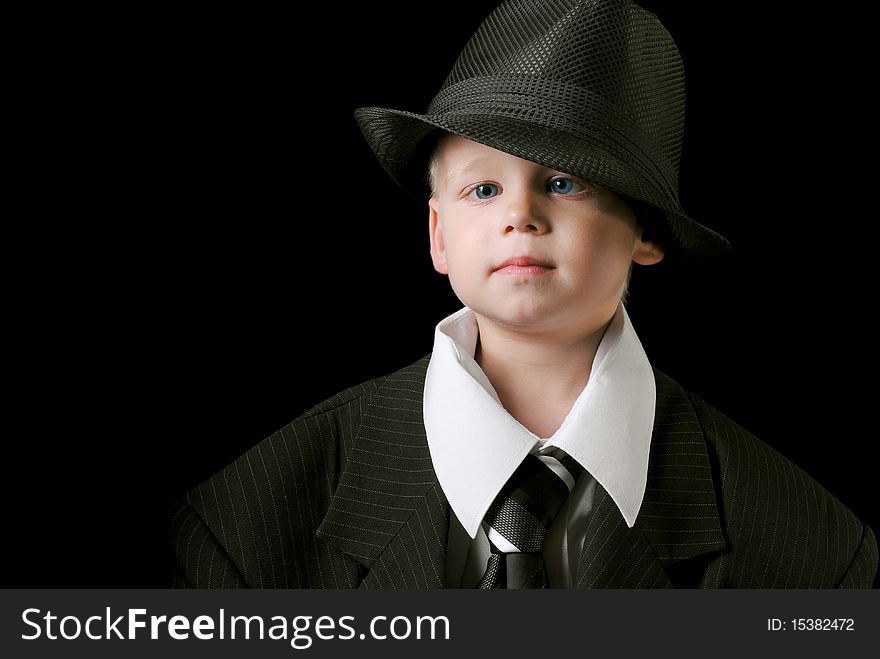 Cute boy with blue eyes wearing adult suit with a tie and a hat isolated on a black background. Cute boy with blue eyes wearing adult suit with a tie and a hat isolated on a black background