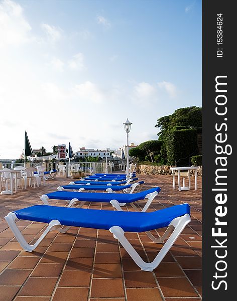A vertical image of a row of blue sun loungers on a terracota tiled patio in early morning sunshine
